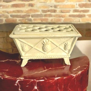 May include: A white cast iron planter with a lattice top and pineapple accents. The planter has four legs and is sitting on a red marble surface.
