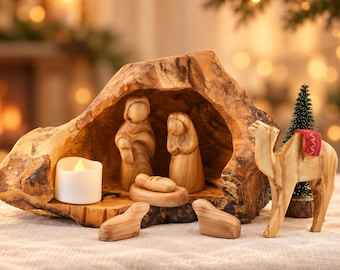 May include: A nativity scene carved from light brown wood, featuring stylized figures of two adults and an infant inside a hollowed log structure. A white LED tea light, two small animal figures, and a camel with a red patterned saddle are included.