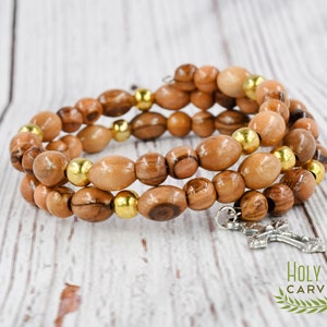 May include: A triple-strand bracelet made of olive wood beads with gold accents and a silver cross charm. The bracelet is on a light wood background.