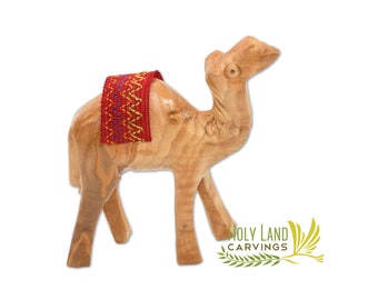 May include: A hand-carved wooden camel figurine with a red and gold patterned saddle. The camel is standing on a white background.