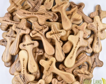 May include: A pile of small wooden crosses with various bible verses engraved on them. The crosses are made of olive wood and have a natural wood finish. The verses include "God is Love", "Jesus Heals", "The Lord is my strength", and "Peace be with you".