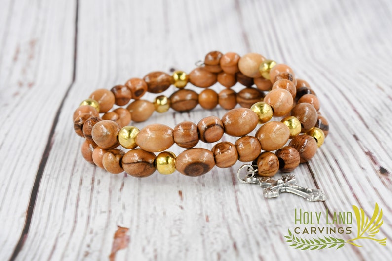 May include: A triple-strand bracelet made of olive wood beads with gold accents and a silver cross charm. The bracelet is on a light wood background.