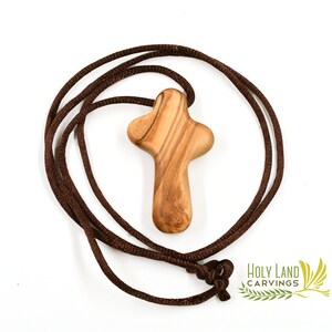 Puede incluir: Un colgante de cruz de madera marr&oacute;n en un collar de cord&oacute;n marr&oacute;n. La cruz est&aacute; tallada en madera de olivo y tiene un dise&ntilde;o simple y tradicional. El collar est&aacute; hecho de un cord&oacute;n suave y c&oacute;modo.