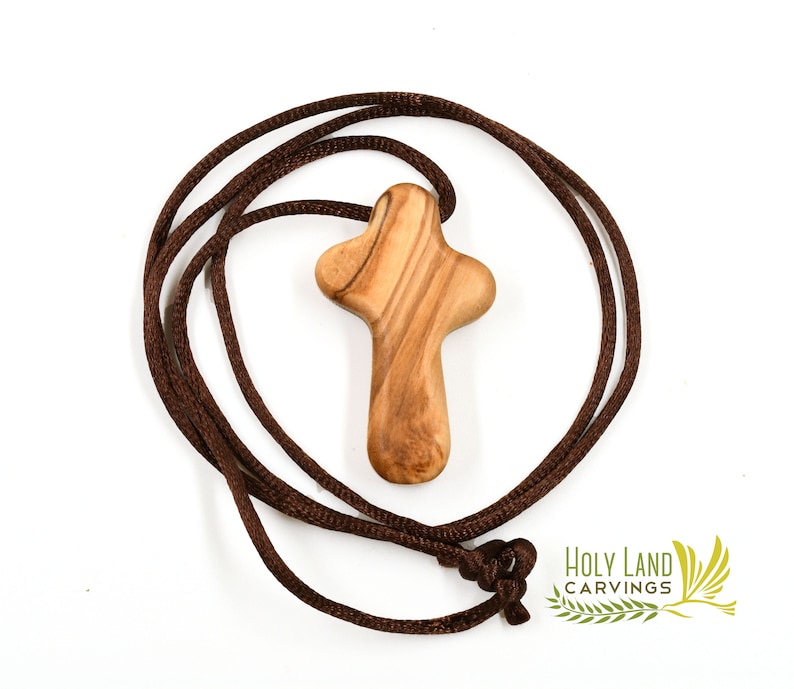 Puede incluir: Un colgante de cruz de madera marr&oacute;n en un collar de cord&oacute;n marr&oacute;n. La cruz est&aacute; tallada en madera de olivo y tiene un dise&ntilde;o simple y tradicional. El collar est&aacute; hecho de un cord&oacute;n suave y c&oacute;modo.