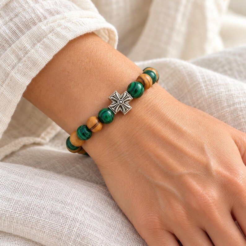 May include: A beaded bracelet featuring alternating round green malachite beads and light brown wooden beads. The bracelet is centered with a small, silver-toned metal cross charm that has an intricate, embossed geometric pattern.