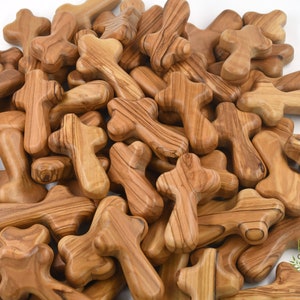 May include: A pile of small, wooden crosses made from olive wood. The crosses are all the same size and shape, and they have a natural, light brown color. The crosses are arranged in a random pattern, and they are all facing the same direction.