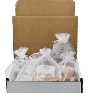 May include: A white cardboard box with a brown cardboard lid. The box is filled with small white organza bags containing wooden olive wood carvings. The box has a label on the bottom that reads "HOLY LAND CARVINGS" with a green olive branch and a white dove.