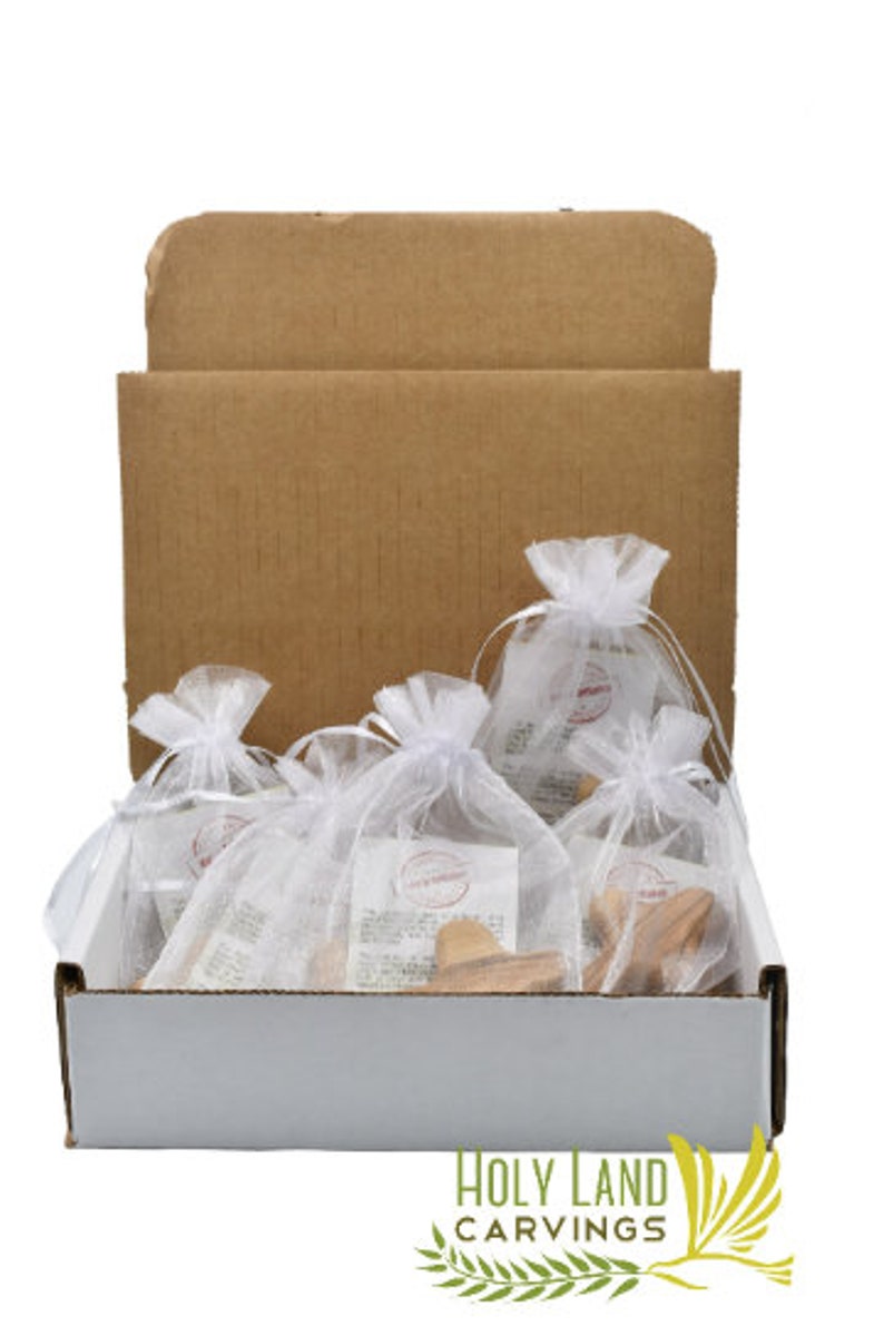 May include: A white cardboard box with a brown cardboard lid. The box is filled with small white organza bags containing wooden olive wood carvings. The box has a label on the bottom that reads "HOLY LAND CARVINGS" with a green olive branch and a white dove.