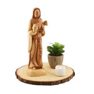 May include: A wooden figurine of a person holding a sheep, standing on a wooden base with a small potted plant and a white candle.