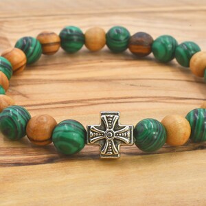 May include: A stretch bracelet featuring alternating round green malachite beads with dark swirling patterns and smooth light brown wooden beads. A central silver-toned metal cross charm with engraved detailing serves as the focal point.