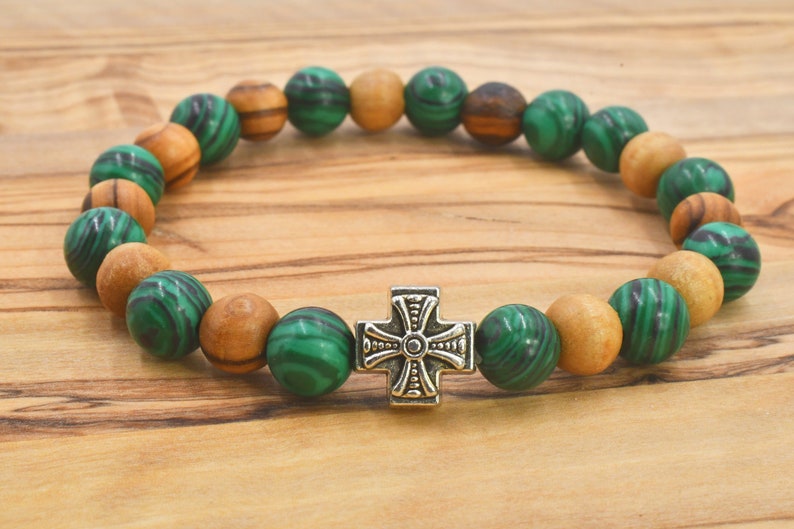 May include: A stretch bracelet featuring alternating round green malachite beads with dark swirling patterns and smooth light brown wooden beads. A central silver-toned metal cross charm with engraved detailing serves as the focal point.