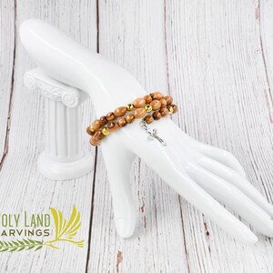 May include: A brown wooden bracelet with gold beads and a silver cross charm. The bracelet is on a white mannequin hand. The background is a white wooden surface.