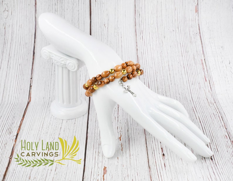 May include: A brown wooden bracelet with gold beads and a silver cross charm. The bracelet is on a white mannequin hand. The background is a white wooden surface.
