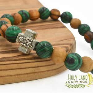May include: A stretch bracelet featuring alternating round green malachite beads and light brown wooden beads. A silver-toned metal cross charm with an embossed figure is centered on the strand. The text Holy Land Carvings is visible.