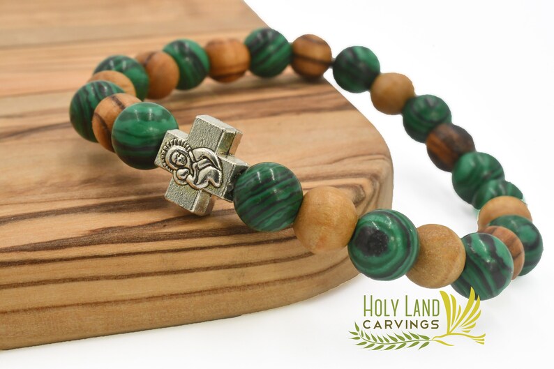 May include: A stretch bracelet featuring alternating round green malachite beads and light brown wooden beads. A silver-toned metal cross charm with an embossed figure is centered on the strand. The text Holy Land Carvings is visible.