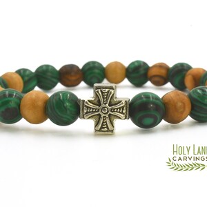 May include: A stretch bracelet featuring alternating round green malachite beads with dark swirling patterns and smooth light brown wooden beads. A central metallic cross charm with an embossed design sits in the middle. The text HOLY LAND CARVINGS is visible in the bottom right corner.