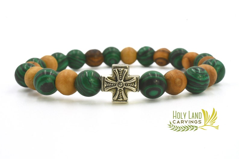 May include: A stretch bracelet featuring alternating round green malachite beads with dark swirling patterns and smooth light brown wooden beads. A central metallic cross charm with an embossed design sits in the middle. The text HOLY LAND CARVINGS is visible in the bottom right corner.