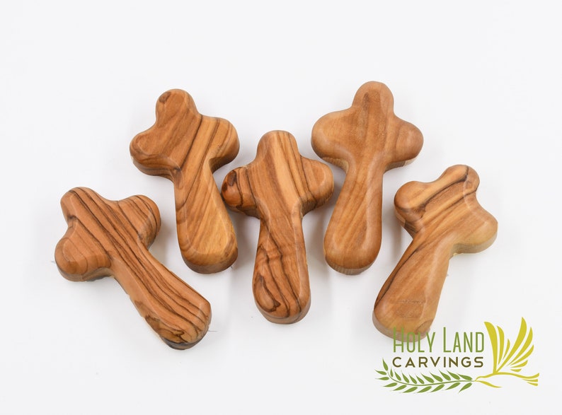 May include: Five small wooden crosses made from olive wood. The crosses are a light brown color and have a natural wood grain. The crosses are all different shapes and sizes.