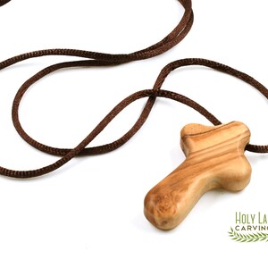 Puede incluir: Un colgante de cruz de madera marr&oacute;n con un cord&oacute;n marr&oacute;n. La cruz est&aacute; hecha de madera de olivo y tiene un dise&ntilde;o simple y r&uacute;stico. El cord&oacute;n mide aproximadamente 50 cm de largo.