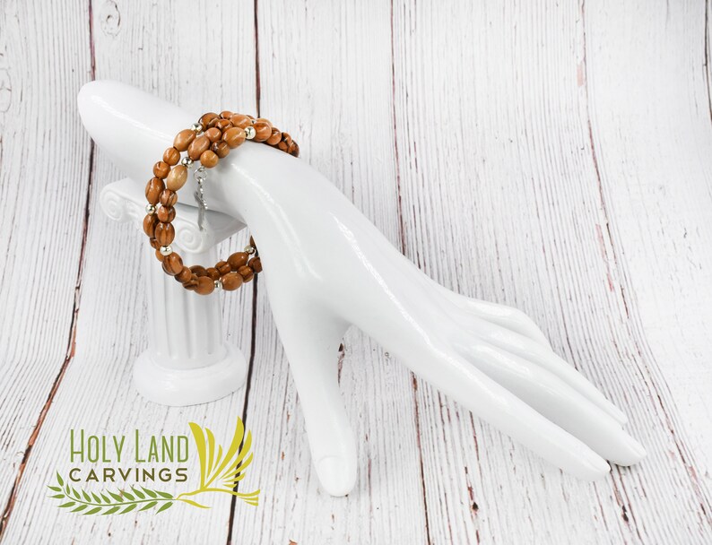 May include: A brown olive wood bracelet with silver beads and a silver charm. The bracelet is on a white mannequin hand. The background is a white wooden surface. The text "HOLY LAND CARVINGS" is visible in the lower left corner of the image.