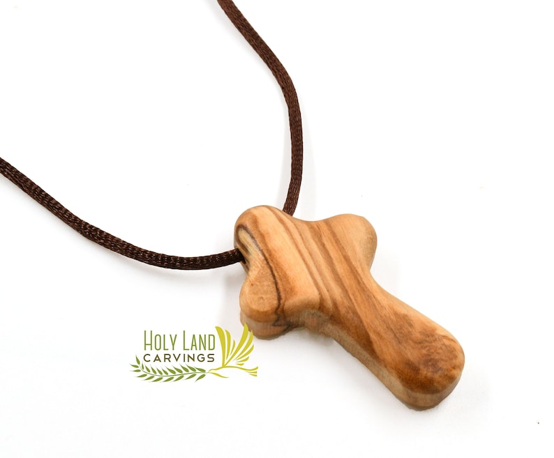 Puede incluir: Un peque&ntilde;o colgante de cruz de madera con un collar de cord&oacute;n marr&oacute;n. La cruz est&aacute; hecha de madera de olivo y tiene un patr&oacute;n de veta de madera natural.