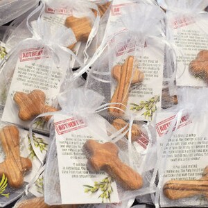 May include: A close-up of several small wooden crosses in white organza bags. Each bag contains a card with the text "AUTHENTIC OLIVE WOOD" and a description of the wood's origin. The crosses are made from olive wood and have a natural wood grain.