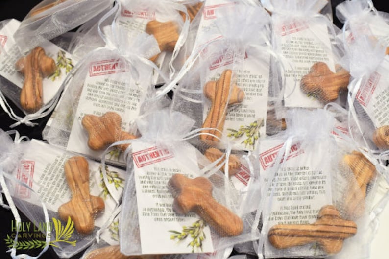 May include: A close-up of several small wooden crosses in white organza bags. Each bag contains a card with the text "AUTHENTIC OLIVE WOOD" and a description of the wood's origin. The crosses are made from olive wood and have a natural wood grain.