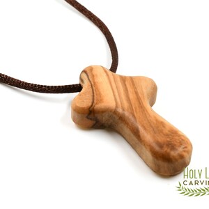 Puede incluir: Un peque&ntilde;o colgante de cruz de madera hecho de madera de olivo. La cruz es de dise&ntilde;o simple y tradicional, con un cord&oacute;n marr&oacute;n.