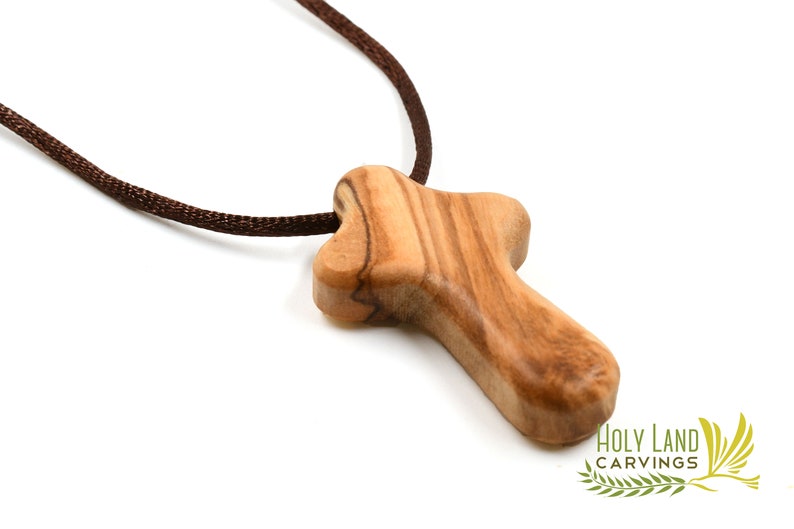Puede incluir: Un peque&ntilde;o colgante de cruz de madera hecho de madera de olivo. La cruz es de dise&ntilde;o simple y tradicional, con un cord&oacute;n marr&oacute;n.