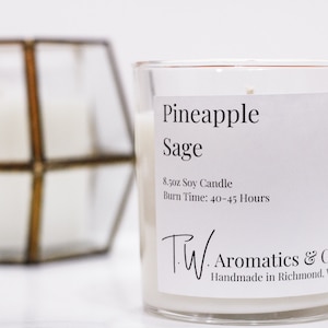 May include: A clear glass jar candle with a white label. The label reads "Pineapple Sage" and "8.5oz Soy Candle, Burn Time: 40-45 Hours". The label also includes the text "TW. Aromatics & Co. Handmade in Richmond, VA".
