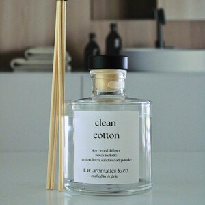 May include: A clear glass reed diffuser bottle with a black cap and a white label reading clean cotton, 4oz reed diffuser, notes include: cotton, linen, sandalwood, powder, t.w. aromatics & co. crafted in virginia, accompanied by light wooden reeds.