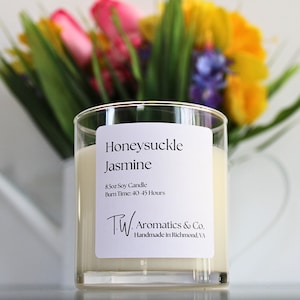 May include: A clear glass jar candle with a white label that reads "Honeysuckle Jasmine 8.5oz Soy Candle Burn Time: 40-45 Hours TW Aromatics & Co. Handmade in Richmond, VA"