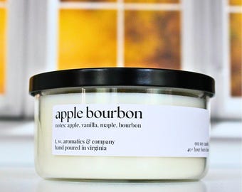 May include: A clear glass jar candle with a black lid. The label reads "apple bourbon" with scent notes listed below. The candle is hand-poured in Virginia and has a 40+ hour burn time.