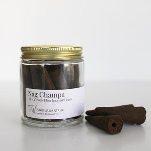 May include: A clear glass jar with a gold lid filled with dark brown incense cones. The label reads "Nag Champa 12-2" Back Flow Incense Cones" and "TW Aromatics & Co. Crafted in Richmond, VA." Three incense cones are next to the jar.