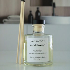 May include: A clear glass reed diffuser bottle with a black cap and a white label reading palo santo + sandalwood, 4oz reed diffuser, notes include: palo santo, patchouli, sandalwood, t.w. aromatics & co. crafted in virginia, with light wooden reeds.