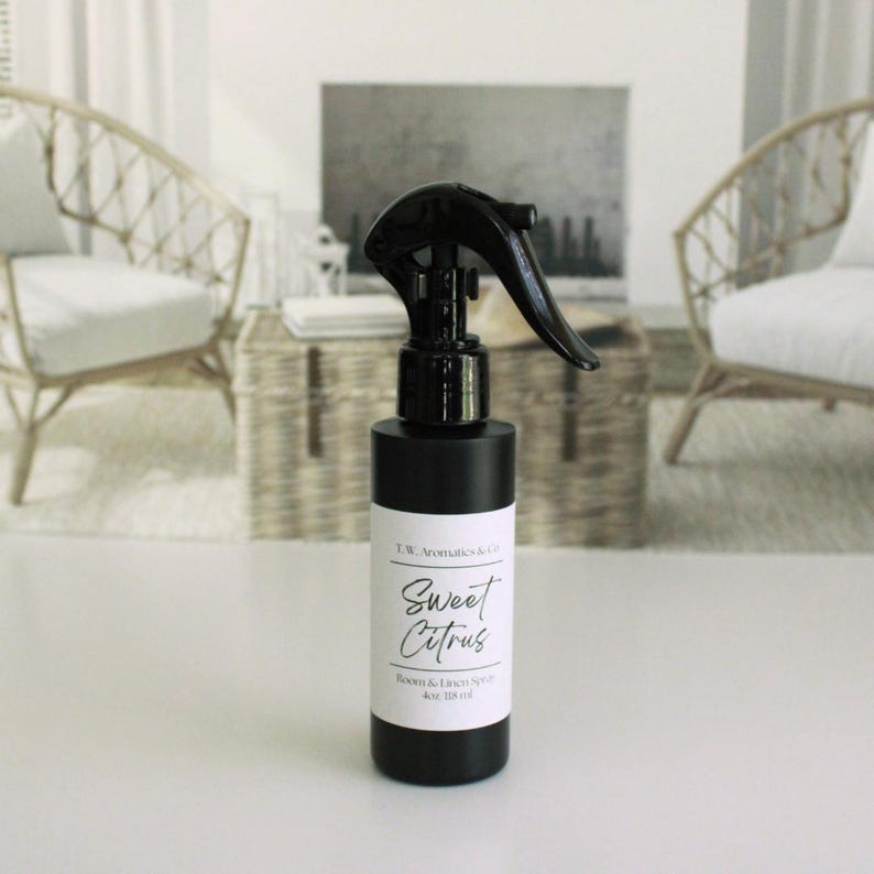 May include: A black room and linen spray bottle with a black trigger. The label reads "Sweet Citrus" and "Room & Linen Spray 4oz 118ml". The bottle is on a white surface, with wicker chairs and a painting in the background.