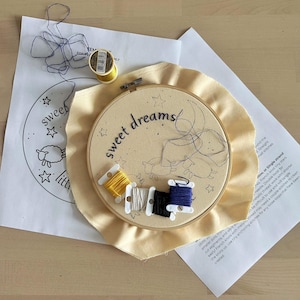 May include: An embroidery hoop containing cream-colored fabric with the words sweet dreams and a sheep outline printed on it. Five small plastic bobbins with yellow, white, black, and blue embroidery thread rest on the fabric. A paper pattern and a spool of yellow thread are nearby.