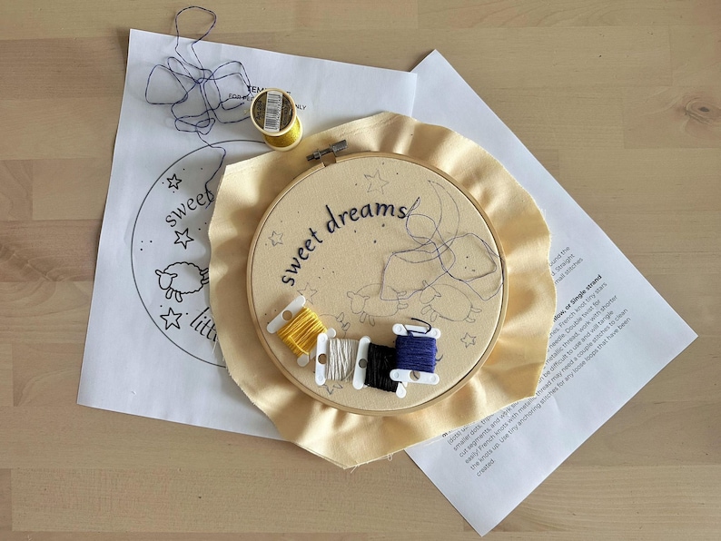 May include: An embroidery hoop containing cream-colored fabric with the words sweet dreams and a sheep outline printed on it. Five small plastic bobbins with yellow, white, black, and blue embroidery thread rest on the fabric. A paper pattern and a spool of yellow thread are nearby.