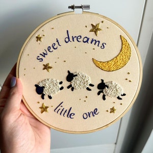 May include: A circular wooden embroidery hoop containing cream-colored fabric with hand-stitched designs. The embroidery features three white sheep with black faces and legs, a yellow crescent moon, gold stars, and the text "sweet dreams little one" in blue.
