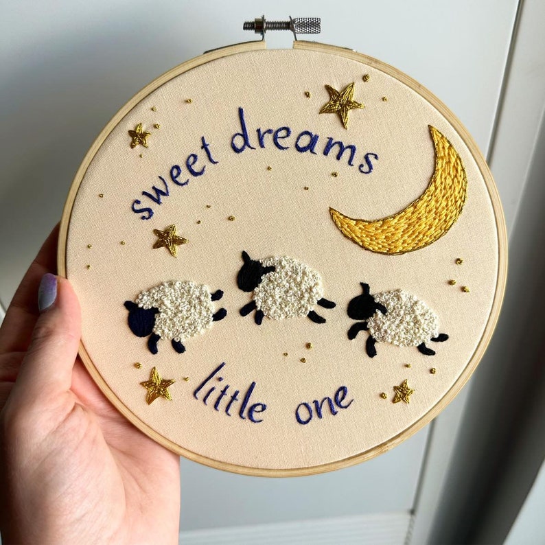 May include: A circular wooden embroidery hoop containing cream-colored fabric with hand-stitched designs. The embroidery features three white sheep with black faces and legs, a yellow crescent moon, gold stars, and the text "sweet dreams little one" in blue.