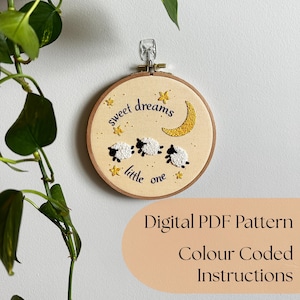 May include: A circular wooden embroidery hoop containing cream-colored fabric with hand-stitched designs of three white sheep, a yellow crescent moon, and yellow stars. The text "sweet dreams little one" is embroidered in dark blue thread.