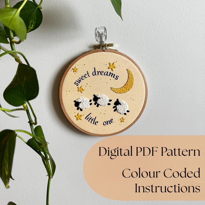 May include: A circular wooden embroidery hoop containing cream-colored fabric with hand-stitched designs of three white sheep, a yellow crescent moon, and yellow stars. The text "sweet dreams little one" is embroidered in dark blue thread.