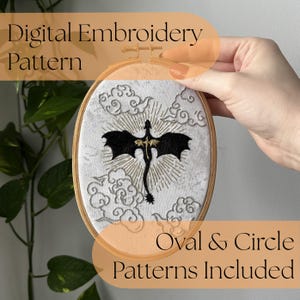 May include: An oval wooden embroidery hoop containing white fabric with a black dragon silhouette stitched in the center. The dragon is surrounded by gold sunburst rays and grey embroidered clouds. Text reads Digital Embroidery Pattern.
