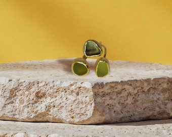 May include: A gold-toned ring featuring three olive-green, irregularly shaped stones. The ring is displayed on a textured, light-colored stone surface against a yellow background. The stones have a polished appearance.