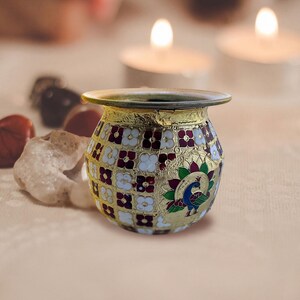 May include: A small, round metal pot featuring a gold-toned finish with an intricate enamel pattern of white and maroon flowers. The vessel includes a prominent peacock motif with green, blue, and pink accents, and a wide, flared circular rim.