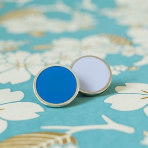 Peut inclure: Deux boucles d'oreilles rondes avec des bordures argentées. L'une est bleu vif, l'autre lilas clair. Elles reposent sur un fond turquoise orné de motifs floraux blancs et dorés, créant une esthétique délicate et élégante.