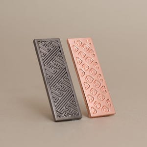 Puede incluir: Dos piezas decorativas rectangulares de metal. Una es de color gris oscuro con un patrón de laberinto geométrico. La otra es de color cobre con un diseño repetido en forma de corazón. Ambas están anguladas sobre un fondo neutro.