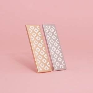 Puede incluir: Dos horquillas rectangulares, una dorada y otra plateada, se alzan una al lado de la otra sobre un fondo rosa. Cada horquilla presenta un patrón geométrico blanco repetido. La horquilla dorada está a la izquierda y la plateada a la derecha.