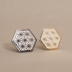 Könnte beinhalten: Zwei sechseckige Pins mit einem weißen geometrischen Muster auf beigefarbenem Hintergrund. Ein Pin ist silber und der andere ist golden.