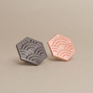 May include: Two hexagonal stud earrings. One is gunmetal gray, the other is rose gold. Each earring features a stylized sun and wave design. The earrings are displayed on a neutral background.